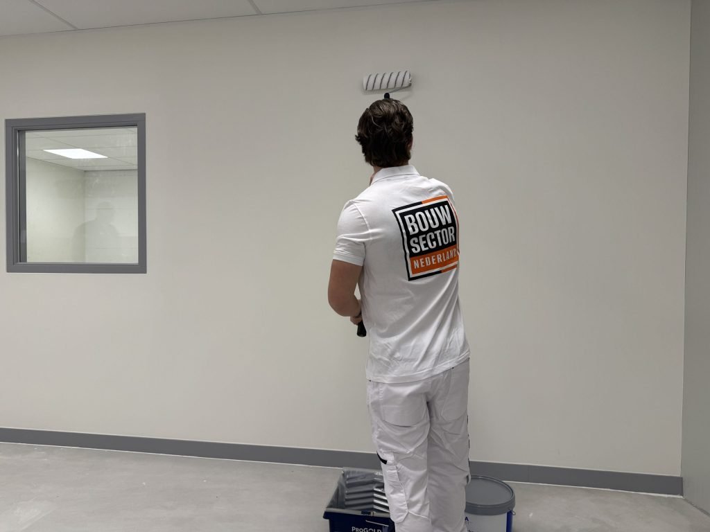 Schilder van Bouwsector Nederland rolt een lichte wand met de verfbak en emmer bij de afgeplakte plint