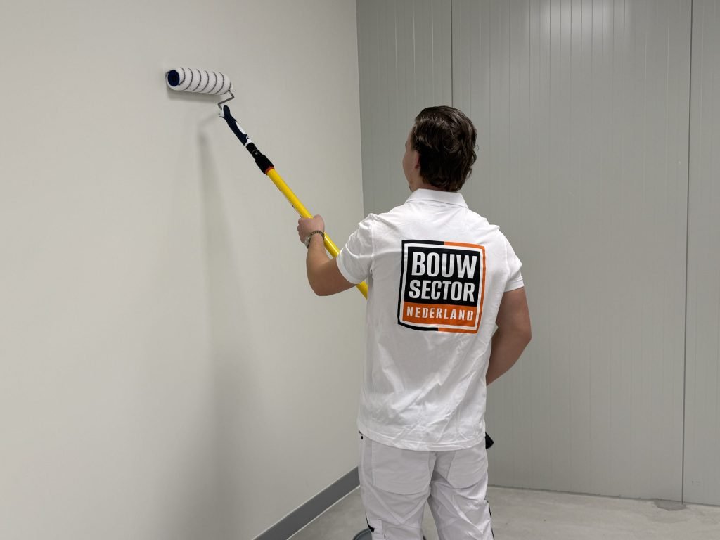 Schilder in witte werkkleding rolt het bovenste deel van een lichte wand met een lange steel