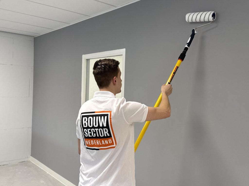 Schilder van Bouwsector Nederland zet een grijze muur in de verf met een roller aan een verlengsteel