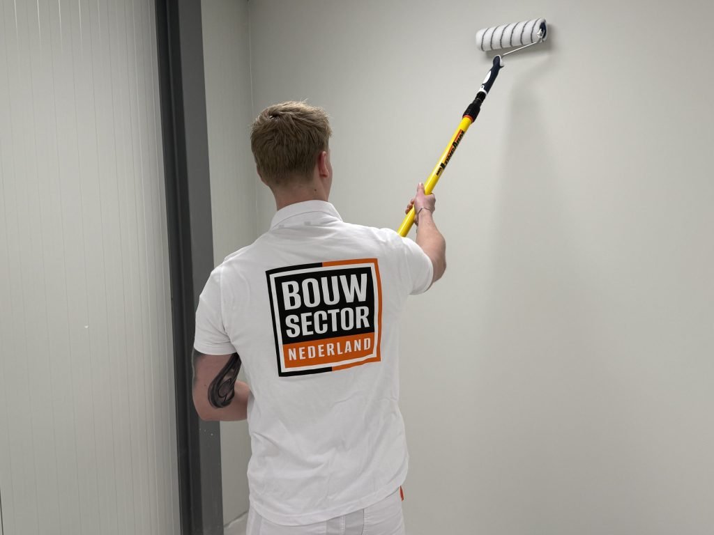 Schilder in bedrijfskleding van Bouwsector Nederland rolt een witte muur met een verlengsteel