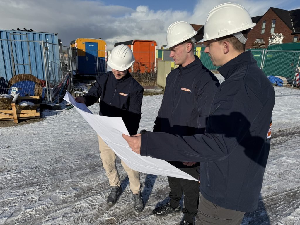 Drie vakmensen van Bouwsector Nederland met witte helm bekijken een bouwtekening op een besneeuwd terrein